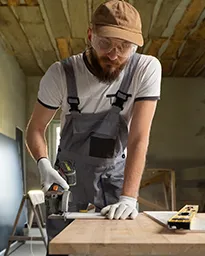 Man in overalls using power tool on wooden plank indoors.