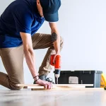 Person kneeling on floor using drill with toolbox nearby.