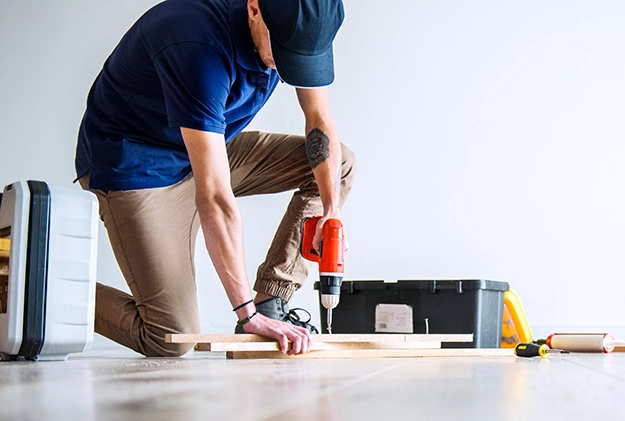 Person kneeling on floor using drill with toolbox nearby.