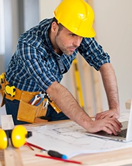 Construction worker in plaid shirt using a laptop with blueprints nearby.
