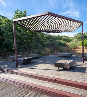 Wooden pergola with benches on a deck, surrounded by trees.