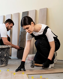 Woman installing wooden flooring with a hammer, man working in background.